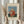 May include: Hands holding a framed art print of Henri Rousseau's "Child with Doll" painting from 1906, showing a young child in a red polka-dot dress holding a small doll against a blue and green background. The print has cream matting with black text above and below the artwork. One hand also holds dried pampas grass stems. Soft natural lighting illuminates the cream-colored curtains in the background.