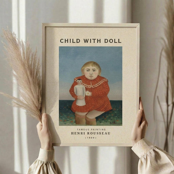 May include: Hands holding a framed art print of Henri Rousseau's "Child with Doll" painting from 1906, showing a young child in a red polka-dot dress holding a small doll against a blue and green background. The print has cream matting with black text above and below the artwork. One hand also holds dried pampas grass stems. Soft natural lighting illuminates the cream-colored curtains in the background.