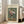 May include: Framed vintage botanical poster featuring colorful flowers in a ribbed glass vase against green background, displayed on light wooden floors next to beige upholstered chair in bright sunlit room with white walls and large windows.