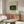 May include: Framed vintage art print featuring tropical jungle scene with banana plants and small figures, displayed above wooden sideboard in modern living room with white curved chair, glass coffee table, and minimalist decor in neutral tones.