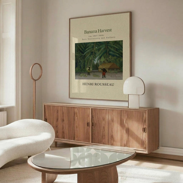 May include: Framed vintage art print featuring tropical jungle scene with banana plants and small figures, displayed above wooden sideboard in modern living room with white curved chair, glass coffee table, and minimalist decor in neutral tones.