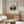 May include: Framed art print featuring a canal scene with bare trees lining waterway under overcast sky, displayed above wooden sideboard with sliding doors. Modern living room setting with cream upholstered chair, glass-top oval coffee table, and white table lamp. Neutral beige walls and natural lighting from window.