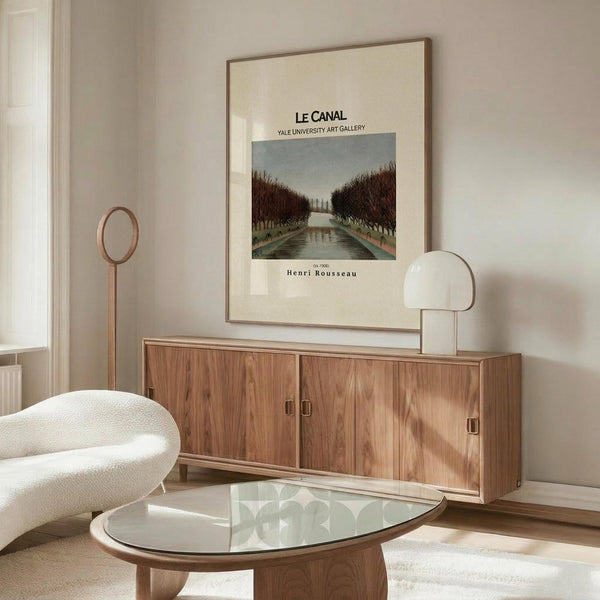 May include: Framed art print featuring a canal scene with bare trees lining waterway under overcast sky, displayed above wooden sideboard with sliding doors. Modern living room setting with cream upholstered chair, glass-top oval coffee table, and white table lamp. Neutral beige walls and natural lighting from window.