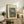 May include: Framed vintage-style botanical print showing colorful flowers in a glass vase, displayed on rustic wood sideboard alongside fern plant, books, and ceramic pottery in neutral-toned living room setting with upholstered furniture