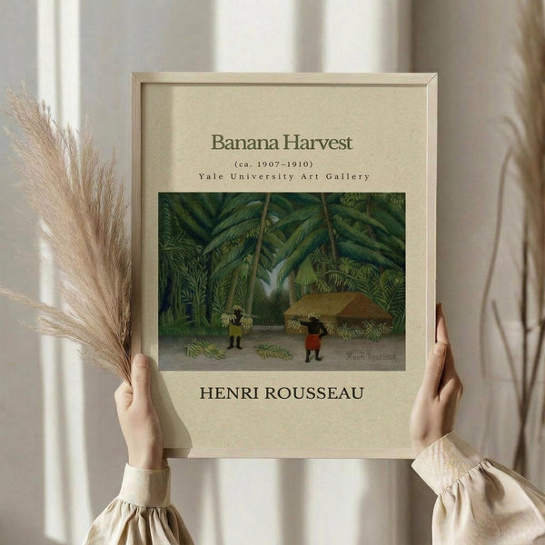 May include: Hands holding a framed vintage art print featuring a tropical jungle scene with banana trees and two small figures harvesting fruit. The print has cream matting with "Banana Harvest (ca. 1907-1910)" and "HENRI ROUSSEAU" text, displayed in a light wood frame. Dried pampas grass visible on the left side against white curtains and neutral interior background.