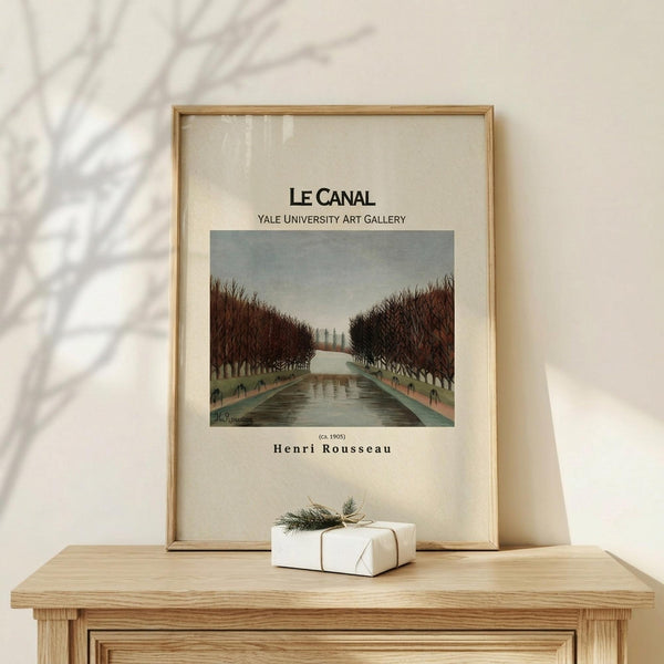 May include: Framed art print of a canal scene with bare trees lining both sides of a reflective waterway under an overcast sky, displayed on a light wood surface alongside a small white wrapped gift with green botanical tie, against a cream wall with plant shadow overlay