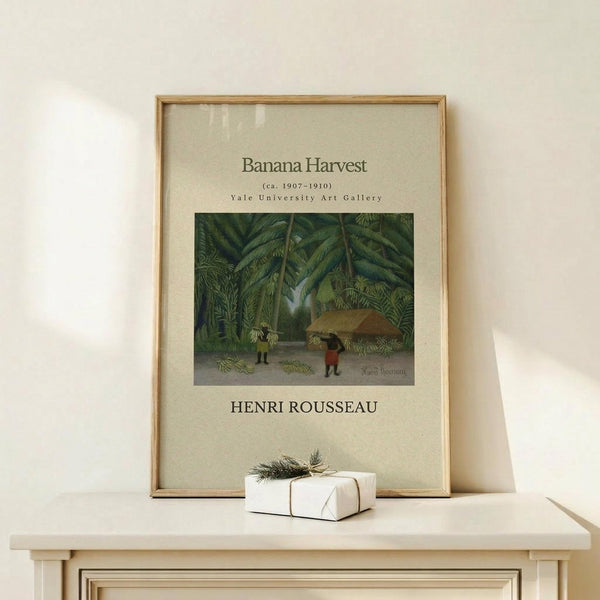 May include: Framed art print displaying Henri Rousseau's tropical jungle painting with lush green foliage and small figures harvesting bananas, presented in natural wood frame with cream matting, positioned on light wooden surface alongside white wrapped gift with twine and small botanical sprig against neutral wall