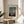 May include: Framed art print displaying a twilight forest scene with bare trees, full moon, and small figures near a cottage. The vintage-style artwork features muted blues and greens in a thin wooden frame with cream matting. Print sits on a round wooden side table next to a beige upholstered chair with decorative pillow, beside a potted plant with long leaves casting shadows on white walls in bright natural lighting.