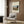 May include: Beige armchair with geometric pillow in modern living room corner, featuring framed vintage art poster of Parisian Seine river scene with Eiffel Tower at sunset. Poster shows bridge, trees, and warm orange sky in naive art style. Gold floor lamp and dark round side table with open book complete the cozy reading nook against wood paneled wall.