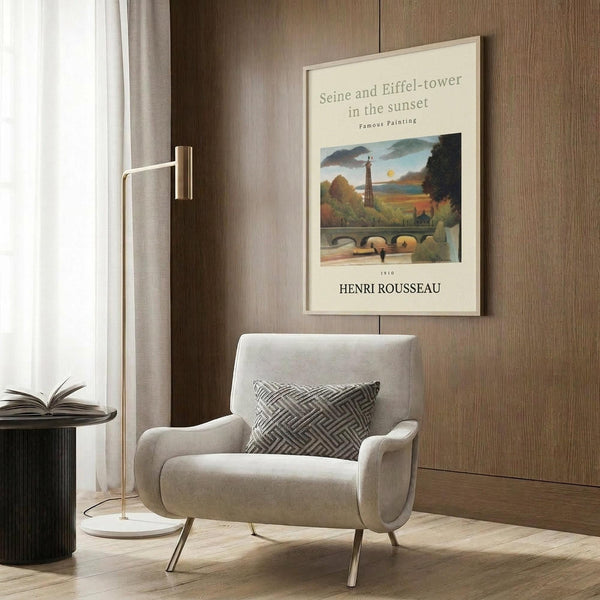 May include: Beige armchair with geometric pillow in modern living room corner, featuring framed vintage art poster of Parisian Seine river scene with Eiffel Tower at sunset. Poster shows bridge, trees, and warm orange sky in naive art style. Gold floor lamp and dark round side table with open book complete the cozy reading nook against wood paneled wall.