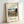 May include: Framed art print leaning against white wall showing Henri Rousseau's 1910 painting of Seine river with Eiffel Tower at sunset. Features naive style landscape with stone bridge, small figures, lush green trees, and warm golden sky. Light wood frame on cream matting with black text identifying artist and date.