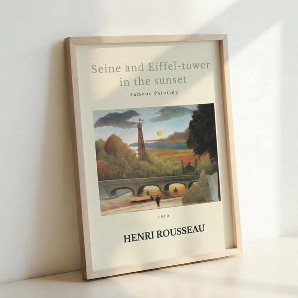 May include: Framed art print leaning against white wall showing Henri Rousseau's 1910 painting of Seine river with Eiffel Tower at sunset. Features naive style landscape with stone bridge, small figures, lush green trees, and warm golden sky. Light wood frame on cream matting with black text identifying artist and date.