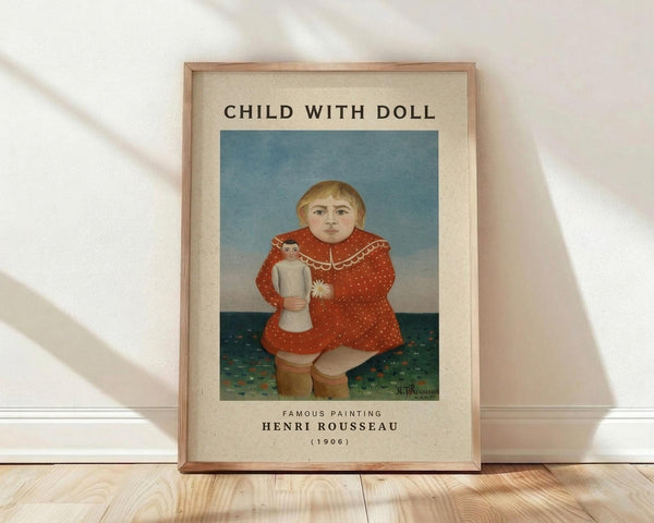 May include: Framed art print of Henri Rousseau's 1906 painting featuring a blonde child in a red polka-dotted dress holding a small doll, set against a blue-green background. The print has cream matting with black text identifying the artwork and artist. Wooden frame leans against white textured wall on light wood flooring with soft natural lighting creating diagonal shadows.