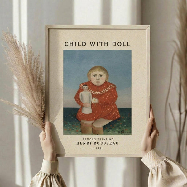 May include: Hands holding a framed art print of Henri Rousseau's "Child with Doll" painting from 1906, showing a blonde child in a red polka dot dress holding a small doll against a blue sky and green ground. The print has a cream mat and wooden frame. One hand also holds dried wheat stalks. Soft natural lighting illuminates the scene against flowing curtains.
