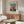 May include: Framed vintage art poster showing a child in red dress holding a white doll, displayed above a walnut wood credenza in a modern living room with cream upholstered chair, glass oval coffee table, and white table lamp. Soft natural lighting creates warm shadows on neutral walls.