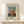 May include: Framed art print of Henri Rousseau's "Child with Doll" painting showing a young child in a red polka dot dress holding a small doll, displayed on a light wood shelf against a cream wall. A small white wrapped gift with evergreen sprig sits beside the natural wood frame.