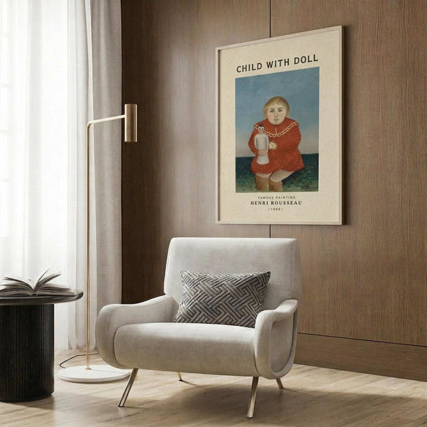 May include: Framed poster print hanging on wood paneled wall above a beige upholstered armchair with geometric pillow. The artwork shows a young child in a red dress holding a doll, painted in a naive folk art style with blue background. Gold floor lamp and dark side table with open book visible nearby.