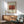 May include: Framed art print of Henri Rousseau's "La Guerre" featuring orange-red tones depicting war scene, displayed above mid-century modern wood credenza with ceramic table lamp, next to sage green upholstered armchair with wooden frame in bright sunlit room with white walls and warm shadows