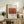 May include: Framed art print displaying Henri Rousseau's "La Guerre" from 1895, featuring orange-red toned jungle scene with figures, styled as National Gallery exhibition poster. Print sits on rustic wooden sideboard alongside fern plant, ceramic vase, lamp, and stacked books in warm neutral living room setting.
