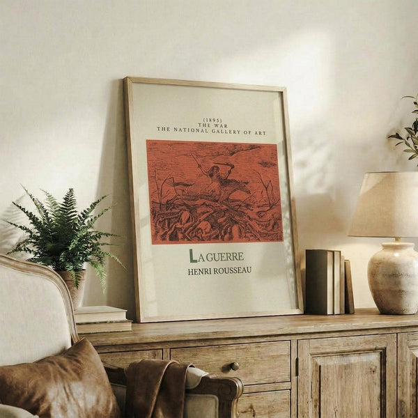 May include: Framed art print displaying Henri Rousseau's "La Guerre" from 1895, featuring orange-red toned jungle scene with figures, styled as National Gallery exhibition poster. Print sits on rustic wooden sideboard alongside fern plant, ceramic vase, lamp, and stacked books in warm neutral living room setting.
