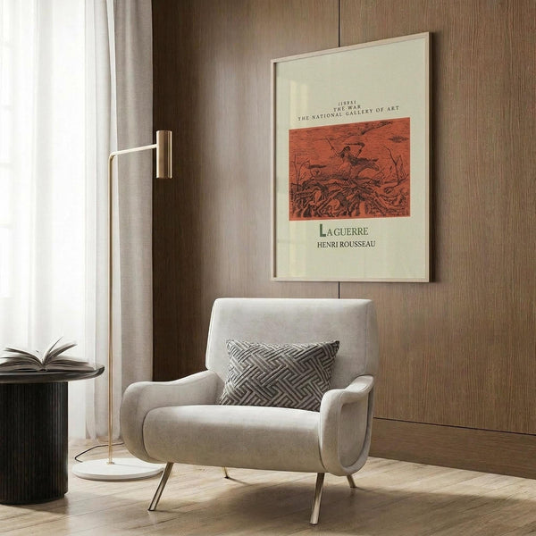 May include: Framed cream-colored poster with reddish-brown war scene artwork displayed on wood-paneled wall above modern beige armchair. Room features brass floor lamp, geometric patterned pillow, dark round side table with open book, and light wood flooring. Soft natural lighting from window with curtains.