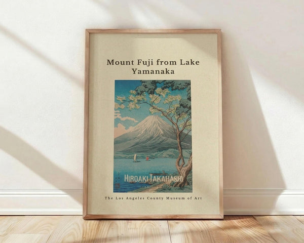 May include: Framed vintage Japanese woodblock print showing Mount Fuji across a lake with cherry blossom tree in foreground, two small boats on blue water, displayed in light wood frame against white textured wall on wooden floor with soft natural lighting creating diagonal shadows