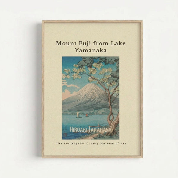 Framed vintage Japanese woodblock print showing Mount Fuji viewed across Lake Yamanaka, with a cherry blossom tree in the foreground and small boats on the blue water. The artwork features soft pastels and traditional ukiyo-e style, displayed in a light wood frame against a cream mat with museum attribution text below.