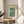 May include: Framed vintage-style Japanese woodblock print showing Mount Fuji across a blue lake with cherry blossom tree in foreground, displayed on light wood round side table next to beige upholstered chair. Natural sunlight creates soft shadows on cream wall. Print features turquoise and pink sunset sky with museum attribution text below artwork in thin wood frame.