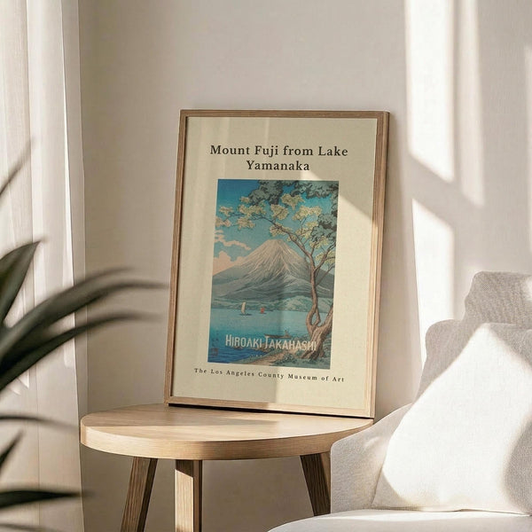 May include: Framed vintage-style Japanese woodblock print showing Mount Fuji across a blue lake with cherry blossom tree in foreground, displayed on light wood round side table next to beige upholstered chair. Natural sunlight creates soft shadows on cream wall. Print features turquoise and pink sunset sky with museum attribution text below artwork in thin wood frame.
