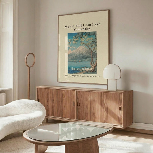 May include: A framed vintage-style Japanese woodblock print poster showing Mount Fuji with a tree and lake in blue and green tones, displayed above a mid-century walnut credenza with sliding doors. The room features a white textured accent chair, glass-top oval coffee table, modern table lamp, and minimalist decor in neutral beige and wood tones.