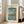 May include: Framed vintage-style Japanese woodblock print poster showing Mount Fuji across Lake Yamanaka with cherry blossom tree in foreground, small boats on blue water, and museum attribution text. Light wood frame leaning against white wall next to beige upholstered chair in bright room with large windows.