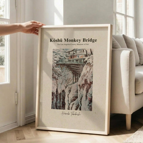 May include: A framed vintage Japanese woodblock print showing a train crossing a wooden bridge through rocky mountain terrain. The artwork features muted earth tones of brown, green, and beige. A person's hand holds the light wood frame against a bright living room with white walls, large windows, and a beige upholstered chair. Natural sunlight creates soft shadows on the hardwood floor.
