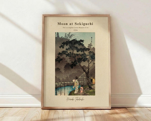 May include: Framed vintage Japanese woodblock print showing evening scene with figures walking near water under large tree, pagoda in background, muted earth tones with blue accents, cream matting in light wood frame, leaning against white wall on wooden floor with natural sunlight shadows