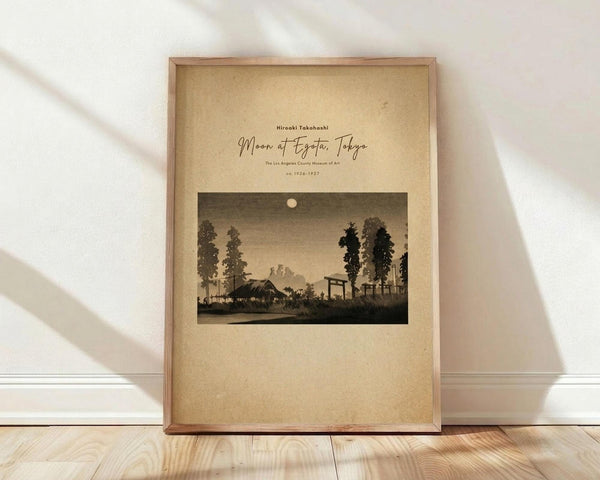 May include: Framed vintage Japanese woodblock print showing a moonlit evening landscape with traditional buildings, tall trees, and torii gate silhouettes against a dusky sky. The artwork has sepia and brown tones on aged paper within a light wood frame, positioned on wooden flooring against a white wall with natural sunlight casting diagonal shadows across the scene.