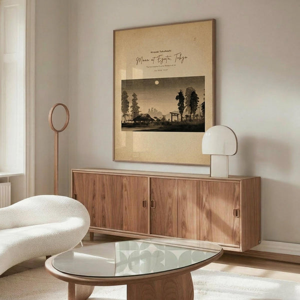 May include: Japanese woodblock print poster displaying moonlit landscape with silhouetted trees and traditional buildings in sepia tones, mounted in thin brown frame on white wall above natural wood credenza. Modern cream curved chair and glass-top oval coffee table visible in minimalist room setting with natural lighting.