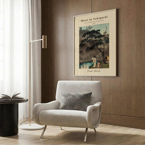 May include: A framed vintage Japanese woodblock print showing figures beneath a large tree by water under a moonlit sky, displayed on wood paneled wall above a cream upholstered armchair with geometric pillow. Modern brass floor lamp and dark round side table with open book complete the neutral-toned reading nook setting.
