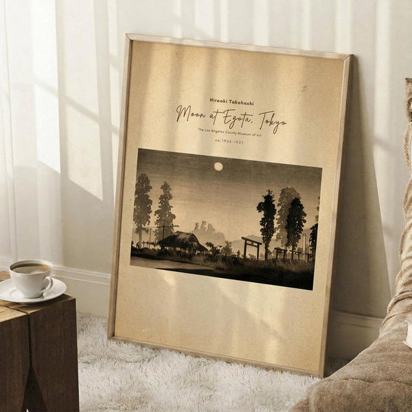 May include: Framed vintage Japanese woodblock print showing moonlit landscape with trees, houses, and small moon in dusky sky, displayed in light wood frame leaning against white wall corner. Coffee cup on wooden block nearby, cream textured rug below, soft natural lighting creating shadows across beige-toned artwork and neutral room setting.