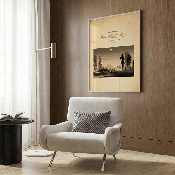 May include: Framed vintage Japanese woodblock print showing moonlit landscape with trees and buildings in earth tones, displayed on wood paneled wall above beige upholstered armchair with geometric patterned pillow, brass floor lamp and side table with open book nearby