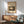 May include: Framed vintage Japanese woodblock print showing moonlit landscape with trees, buildings and mountain silhouettes in sepia tones, displayed above mid-century modern wooden credenza with white ceramic lamp, next to sage green upholstered armchair with wooden frame in sunlit room with white walls