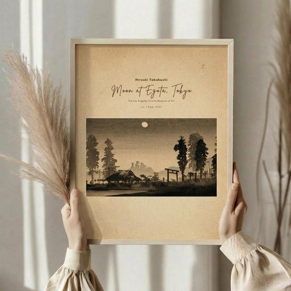 May include: Hands holding a framed vintage Japanese woodblock print showing a moonlit rural landscape with silhouetted trees, traditional buildings, and a torii gate under a sepia-toned sky. The artwork is displayed in a light wooden frame against cream matting, with dried pampas grass visible to the left and soft natural window lighting in the background.