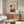May include: Japanese woodblock print poster displayed in modern living room with cream walls, wooden sideboard, white curved chair, glass coffee table, and floor lamp. The vintage artwork features figures in traditional dress near rickshaws with warm coral and teal colors in an aged cream matted frame.
