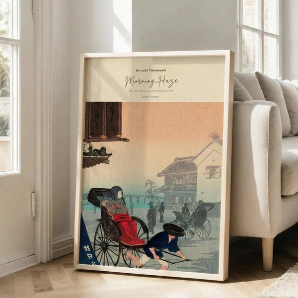 May include: A framed vintage Japanese woodblock print reproduction leaning against a white wall next to a beige upholstered sofa. The artwork shows a morning street scene with a woman in red kimono sitting in a rickshaw, a man in blue crouched beside it, and misty buildings in warm peachy-orange and blue-green tones. Light wood frame, cream matting, displayed on light wooden floors near tall windows with natural lighting.