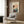 May include: Japanese woodblock print poster displayed in modern reading nook with beige armchair and geometric pillow. Artwork shows traditional scene with rickshaw, figures in colorful robes, and misty landscape in soft peach, teal, and navy tones. Gold floor lamp and dark round side table with open book complete the sophisticated interior setting with wood paneled walls.