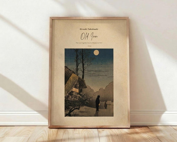 May include: Framed vintage Japanese woodblock print showing a moonlit winter scene with traditional thatched-roof houses, bare trees, and silhouetted figures walking on a snowy path. The artwork features muted blues, grays, and warm earth tones with a cream-colored mat border in a natural wood frame, displayed against white walls on light wooden flooring with soft natural lighting.
