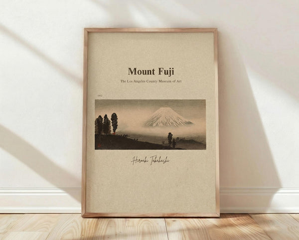 May include: Framed poster print featuring Mount Fuji landscape artwork in muted earth tones with trees in foreground, displayed in light wood frame against white wall on wooden floor with soft natural lighting and shadows