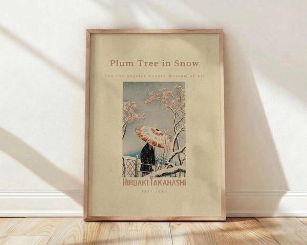 May include: Framed vintage Japanese woodblock print poster leaning against white wall on light wooden floor. The artwork shows a snowy winter scene with a plum tree's delicate pink blossoms, a figure with a red and white striped umbrella near a traditional building structure. Muted gray sky with falling snow creates a serene atmosphere. The print has a cream-colored background with brown text and is displayed in a simple wooden frame with natural wood grain visible.