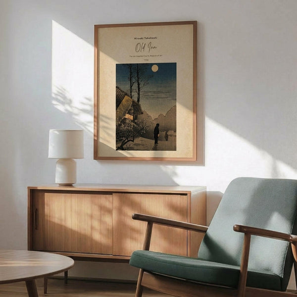 May include: Framed vintage Japanese woodblock print showing a snow-covered village scene with thatched roof buildings, bare trees, and figures walking under a full moon in twilight sky. Print displayed above mid-century modern wooden sideboard with table lamp, next to sage green upholstered armchair with wooden frame. Warm natural lighting creates geometric shadows on white wall.