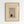 May include: Framed museum poster print mounted on white wall showing Japanese woodblock artwork of figure with red and white umbrella walking past blooming plum tree branches in falling snow. Warm wood frame surrounds cream-colored poster with vintage exhibition styling and artist name at bottom.