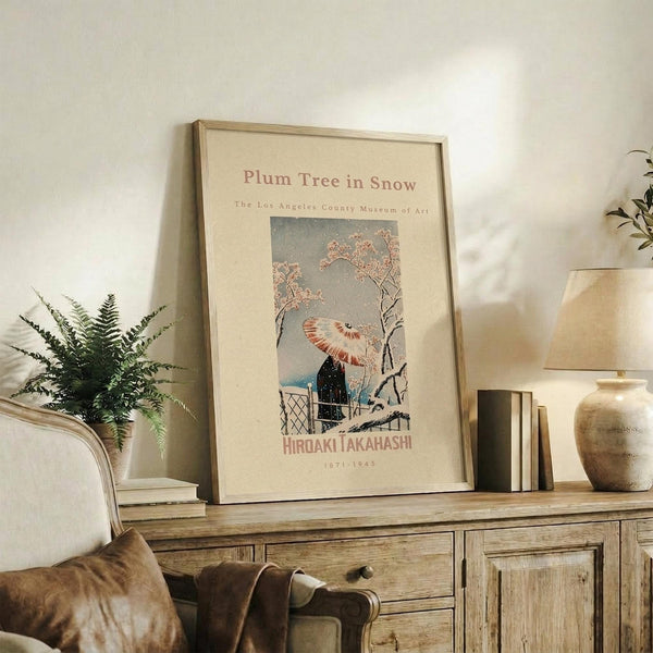May include: Framed vintage Japanese art print featuring a plum tree with pink blossoms in falling snow, with a figure holding a red umbrella beneath bare branches. The cream-matted artwork is displayed on a rustic wooden console alongside a fern plant, brown leather chair, stacked books, and ceramic pottery in a warm, neutral-toned living room setting.