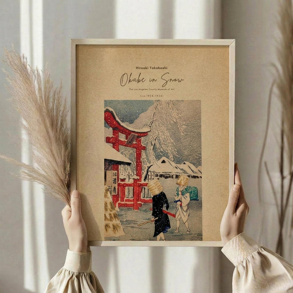 May include: Hands holding a framed vintage Japanese woodblock print showing people with umbrellas walking in snow near a red torii gate and traditional buildings. The artwork has muted colors with pops of red and teal against a tan background. Dried pampas grass sits nearby and white curtains create soft lighting in the background.
